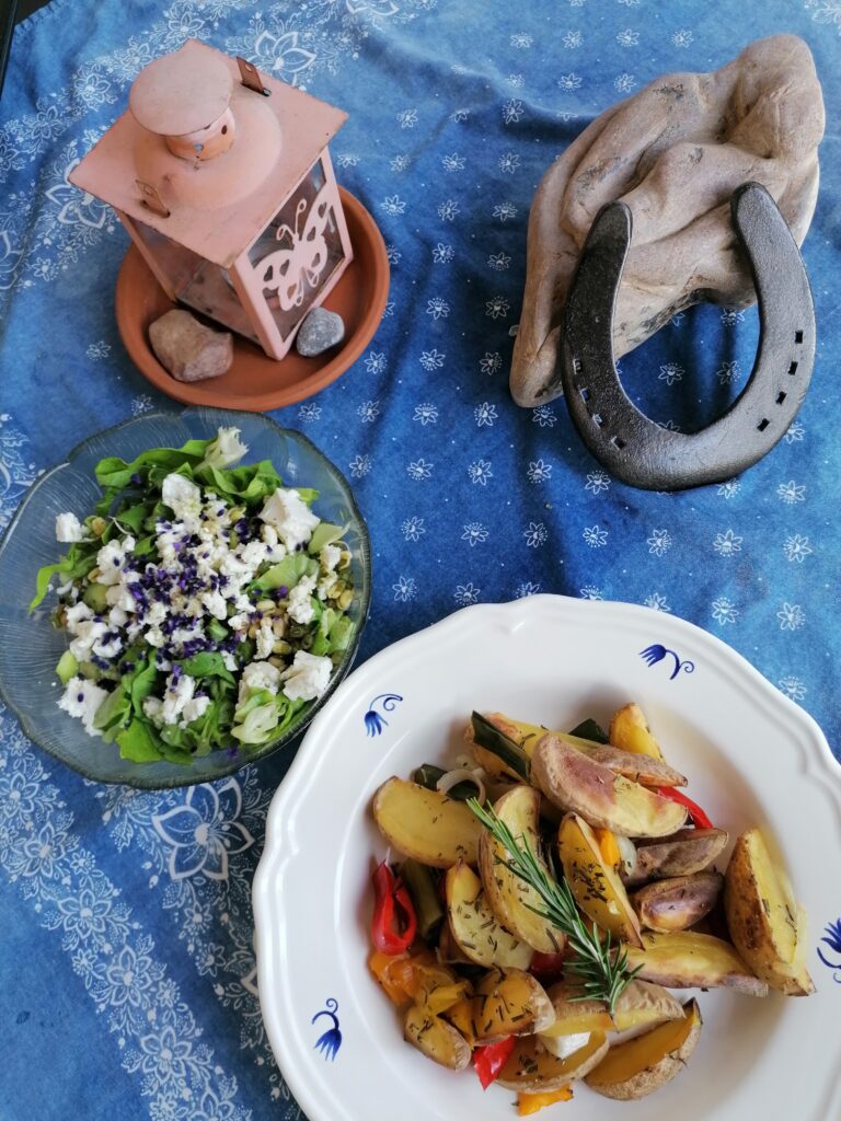 Man sieht einen blaues Tischtuch auf dem eine Laterne und ein Element aus Stein mit Hufeisen steht, daneben eine Schüssel bunter Salat und Ofengemüse mit Rosmarin