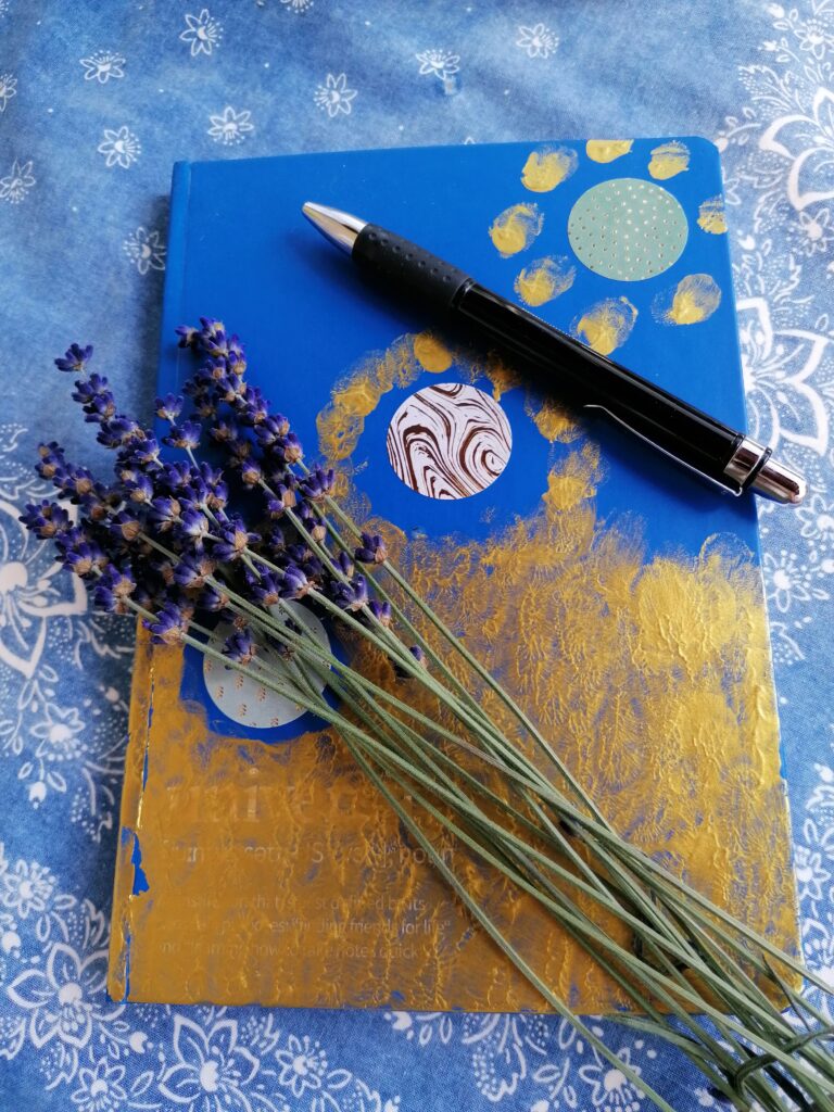 Blau-Goldenes Tagesbuch mit einem Lavendelstrauss obendrauf auf einer blauen Tischdecke